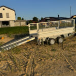 Offener zweiachsiger Kippanhänger mit aufgeklappter Laderampe, abgestellt auf einer sandigen Baustelle.