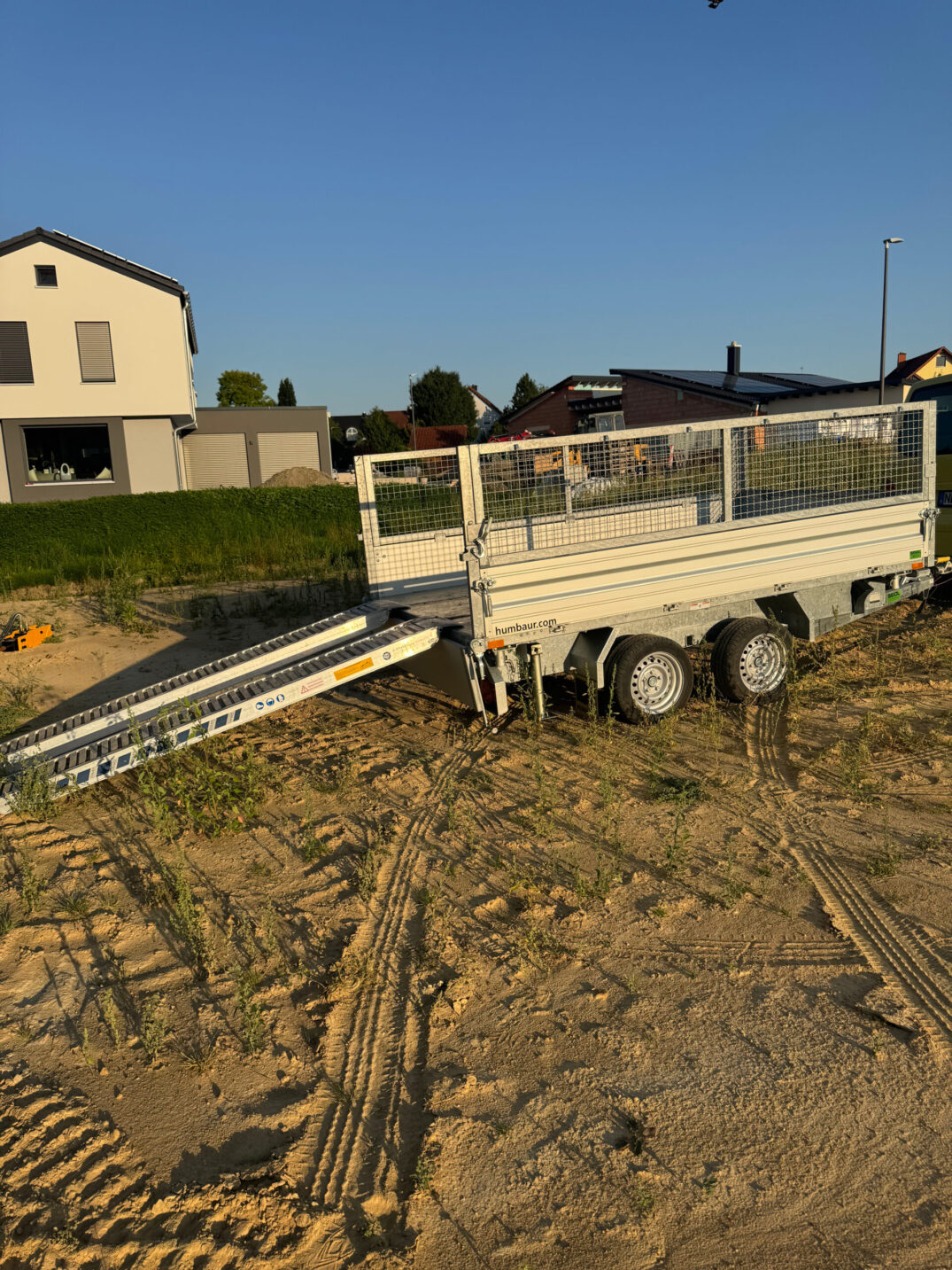 Offener zweiachsiger Kippanhänger mit aufgeklappter Laderampe, abgestellt auf einer sandigen Baustelle.