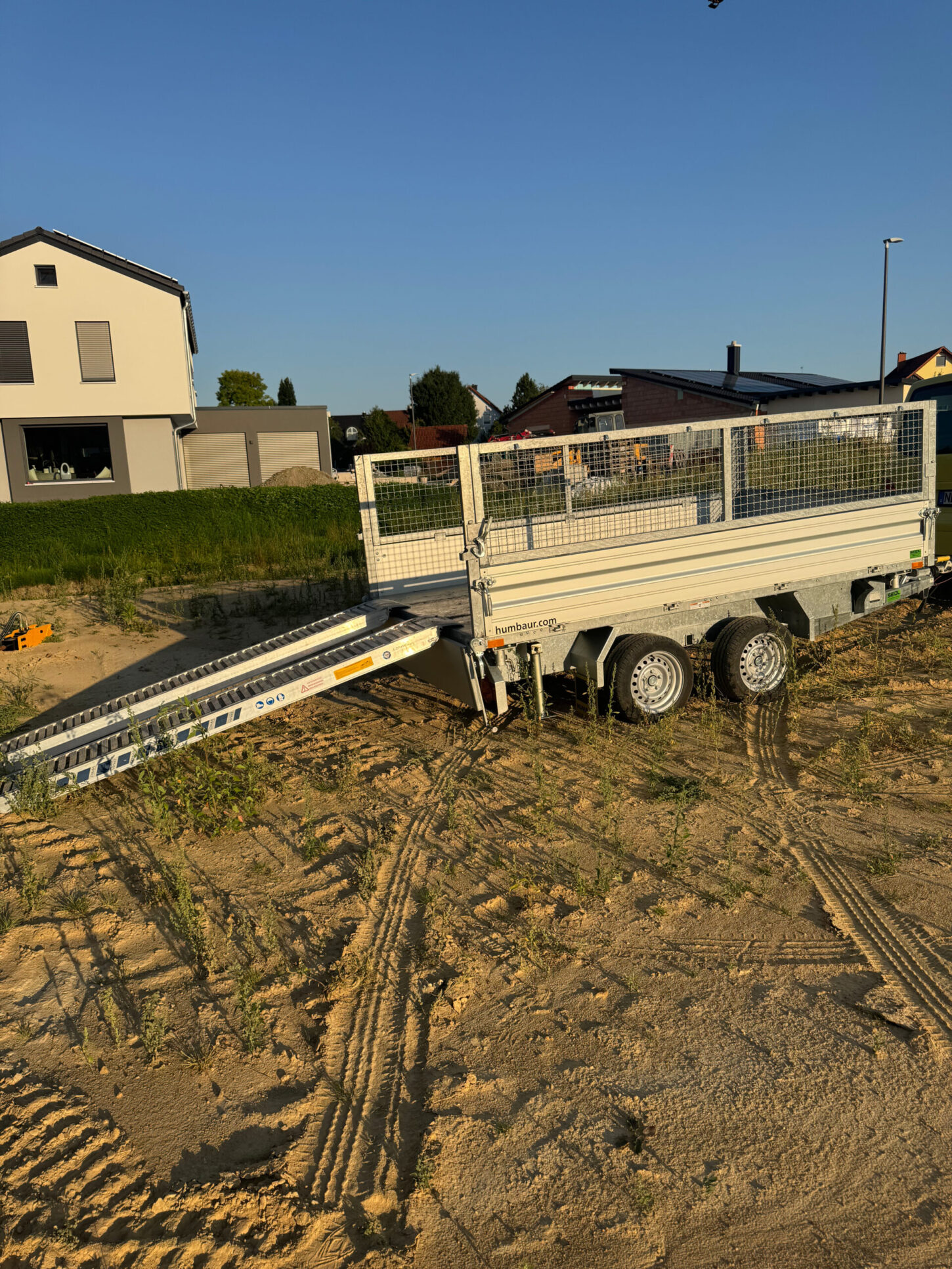 Offener zweiachsiger Kippanhänger mit aufgeklappter Laderampe, abgestellt auf einer sandigen Baustelle.