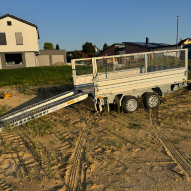Offener zweiachsiger Kippanhänger mit aufgeklappter Laderampe, abgestellt auf einer sandigen Baustelle.