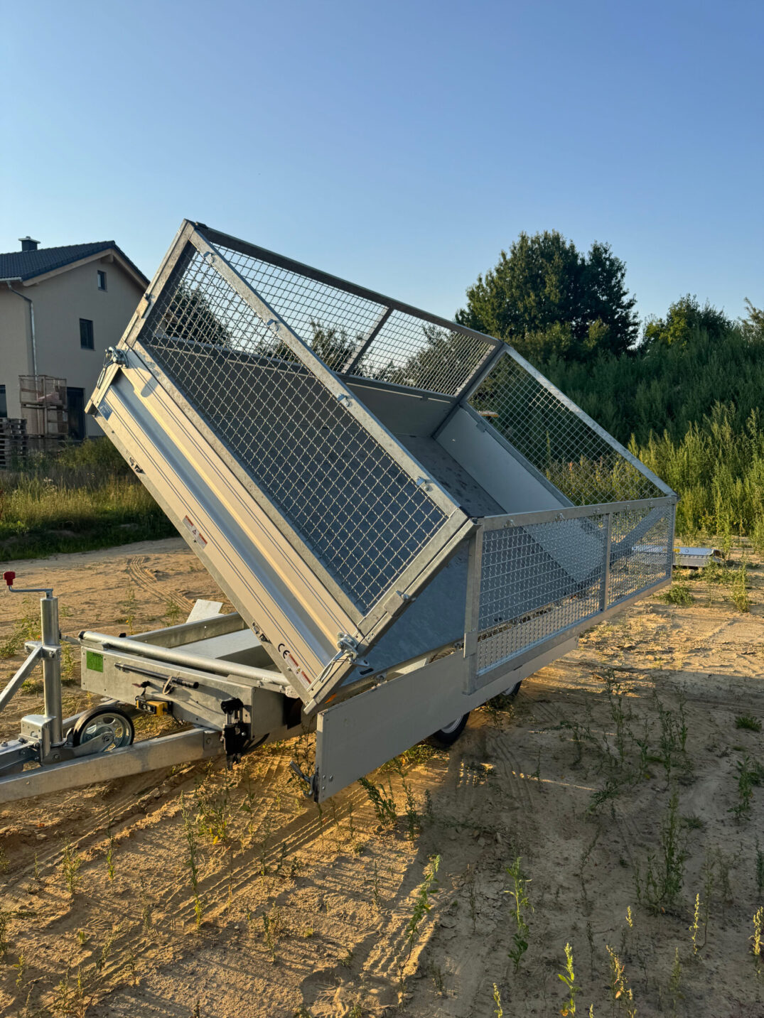 Kippanhänger mit Gitteraufsatz, Ladefläche nach hinten gekippt, abgestellt auf sandigem Untergrund vor Bäumen und Wohnhäusern.
