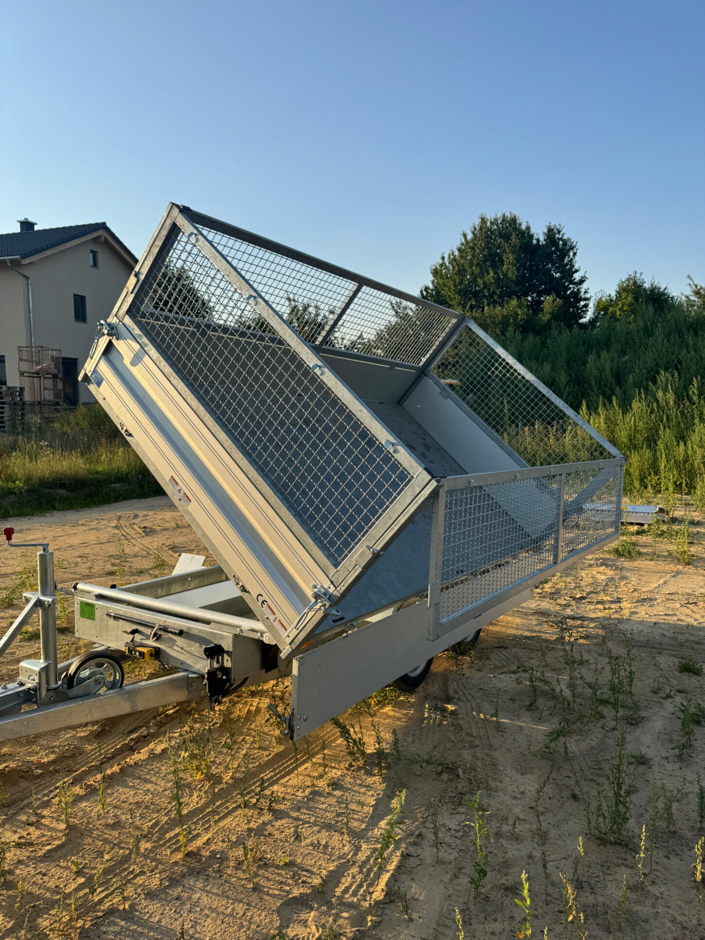 Kippanhänger mit Gitteraufsatz, Ladefläche nach hinten gekippt, abgestellt auf sandigem Untergrund vor Bäumen und Wohnhäusern.