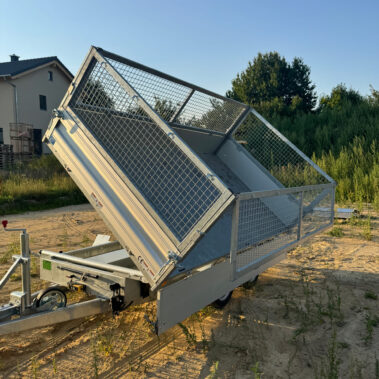 Kippanhänger mit Gitteraufsatz, Ladefläche nach hinten gekippt, abgestellt auf sandigem Untergrund vor Bäumen und Wohnhäusern.