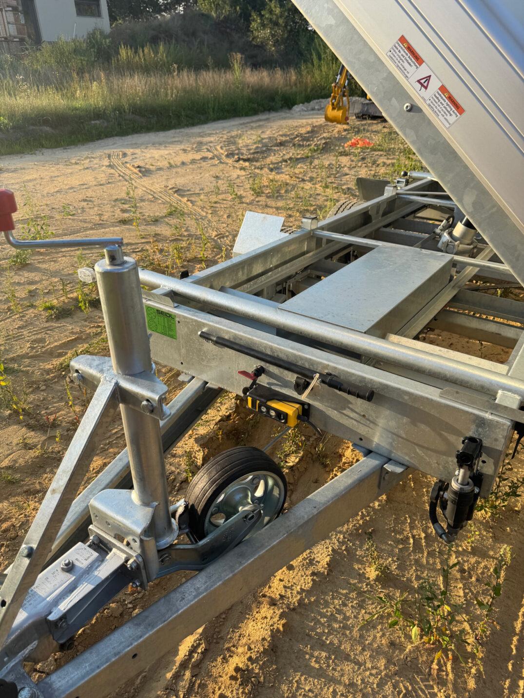 Nahaufnahme der Deichsel eines Kippanhängers mit Stützrad, Kurbel und gelbem Bedienelement, im Hintergrund ein Minibagger.