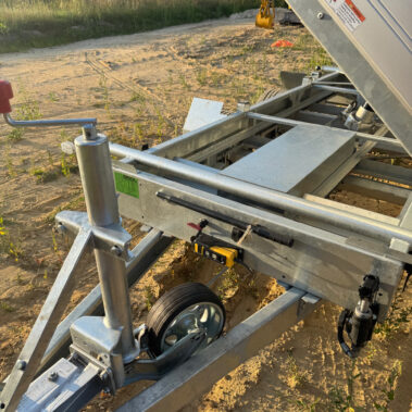 Nahaufnahme der Deichsel eines Kippanhängers mit Stützrad, Kurbel und gelbem Bedienelement, im Hintergrund ein Minibagger.