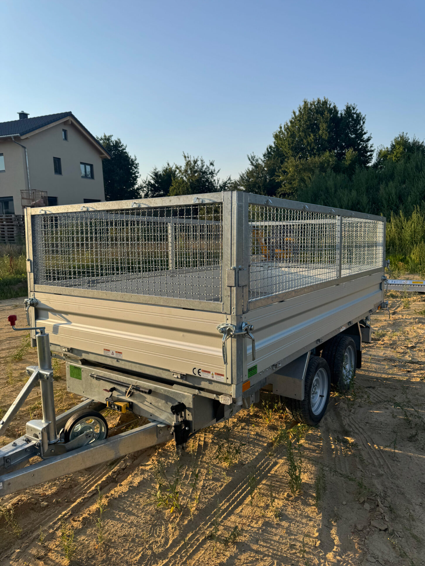 Geschlossener Kippanhänger mit Gitteraufsatz, Doppelachse und Stützrad, geparkt auf einer unbefestigten, sonnigen Fläche.