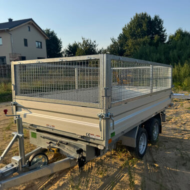 Geschlossener Kippanhänger mit Gitteraufsatz, Doppelachse und Stützrad, geparkt auf einer unbefestigten, sonnigen Fläche.