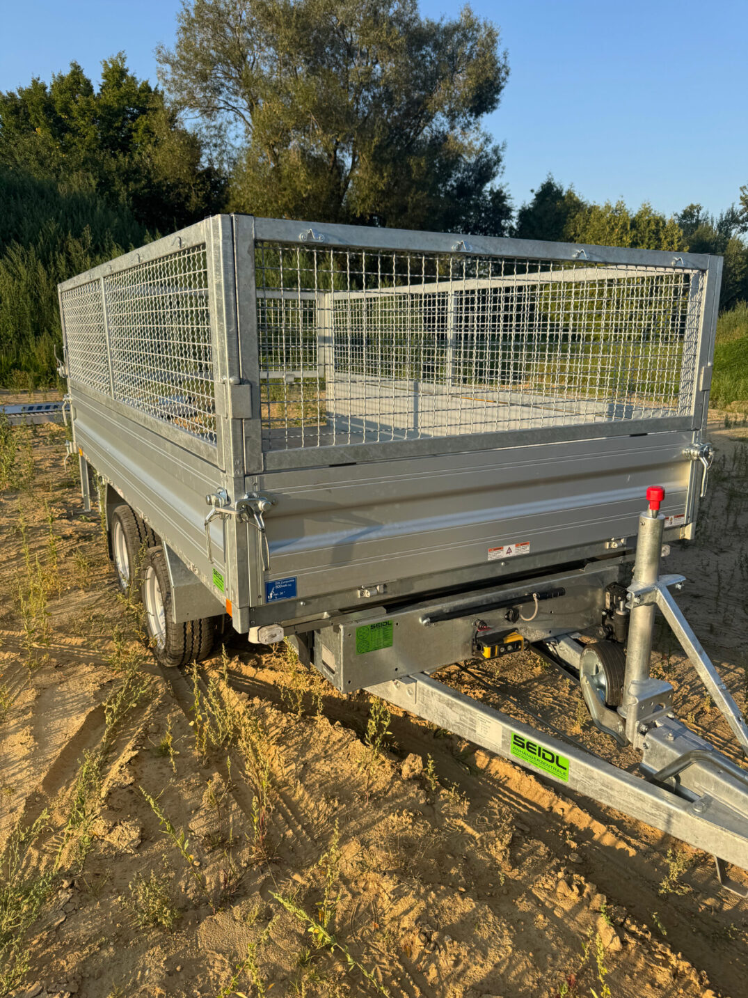 Vorderansicht eines geschlossenen Kippanhängers mit Gitteraufsätzen, abgestützt mit Deichselstütze, im Sonnenlicht.