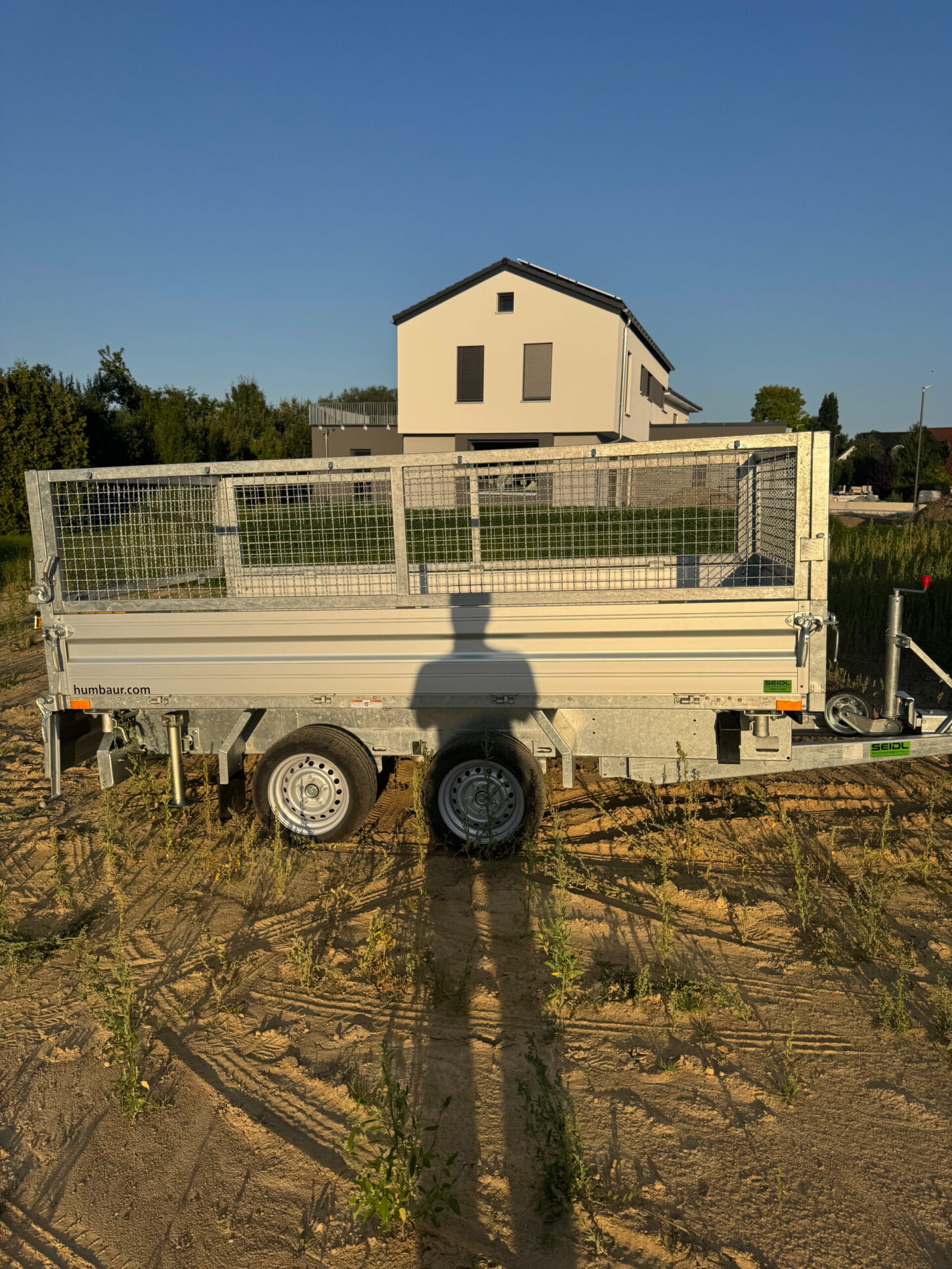 Seitenansicht eines geschlossenen Kippanhängers mit Doppelachse, auf einer sandigen Fläche, Schatten einer Person im Vordergrund.