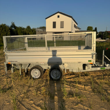 Seitenansicht eines geschlossenen Kippanhängers mit Doppelachse, auf einer sandigen Fläche, Schatten einer Person im Vordergrund.