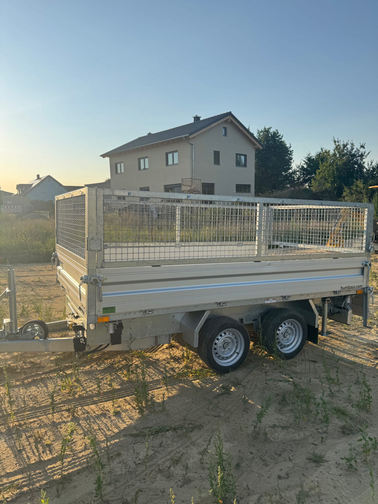 Schrägansicht eines geschlossenen Kippanhängers mit Gitteraufsatz, geparkt auf einer unbefestigten Fläche.
