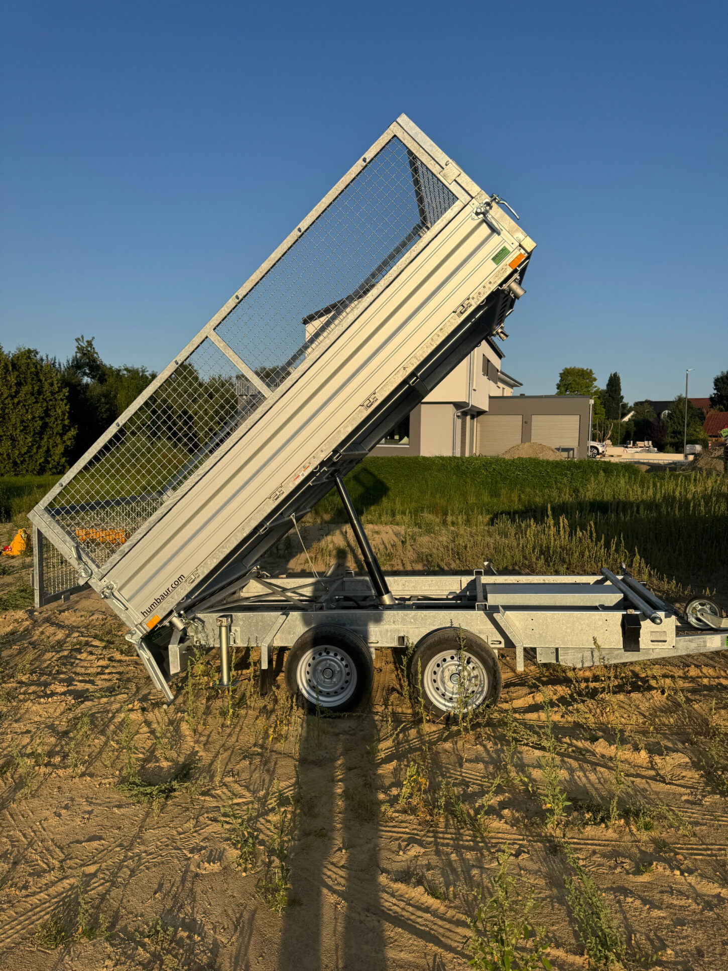 Seitenansicht eines silbernen Kippanhängers mit Gitteraufsätzen, dessen Ladefläche steil angehoben ist, geparkt auf einer sandigen Baustelle bei tiefstehender Abendsonne.