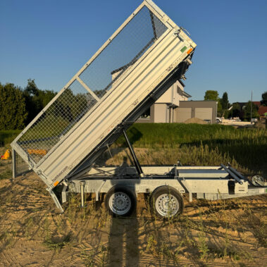 Seitenansicht eines silbernen Kippanhängers mit Gitteraufsätzen, dessen Ladefläche steil angehoben ist, geparkt auf einer sandigen Baustelle bei tiefstehender Abendsonne.