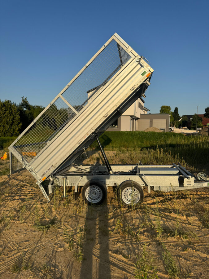 Seitenansicht eines silbernen Kippanhängers mit Gitteraufsätzen, dessen Ladefläche steil angehoben ist, geparkt auf einer sandigen Baustelle bei tiefstehender Abendsonne.