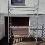 Aufgebautes Rollgerüst aus Aluminium mit Arbeitsplattform und seitlichen Streben, positioniert vor einem geschlossenen Garagentor unter einem Fenster.