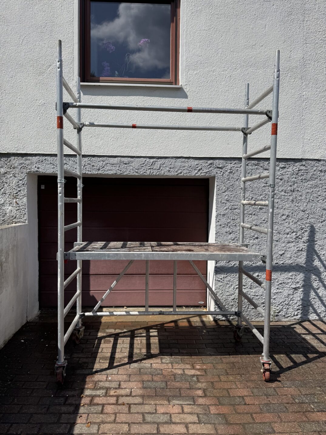 Zerlegtes Rollgerüst mit Aluminiumteilen, darunter eine Plattform und zwei seitliche Stangen, stehend vor einem geschlossenen Garagentor.
