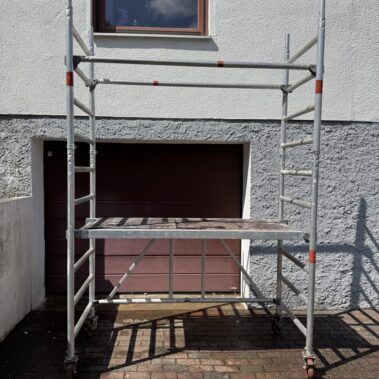 Zerlegtes Rollgerüst mit Aluminiumteilen, darunter eine Plattform und zwei seitliche Stangen, stehend vor einem geschlossenen Garagentor.