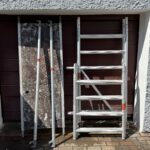 Zerlegtes Rollgerüst mit Aluminiumteilen, darunter eine Plattform und zwei seitliche Stangen, stehend vor einem geschlossenen Garagentor.