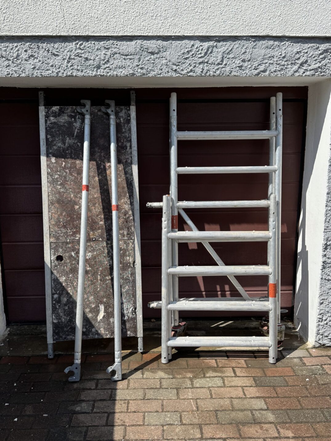 Zerlegtes Rollgerüst mit Aluminiumteilen, darunter eine Plattform und zwei seitliche Stangen, stehend vor einem geschlossenen Garagentor.