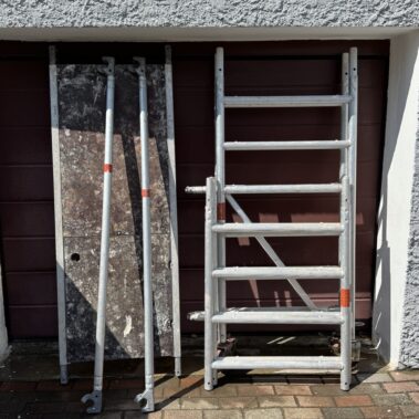 Zerlegtes Rollgerüst mit Aluminiumteilen, darunter eine Plattform und zwei seitliche Stangen, stehend vor einem geschlossenen Garagentor.