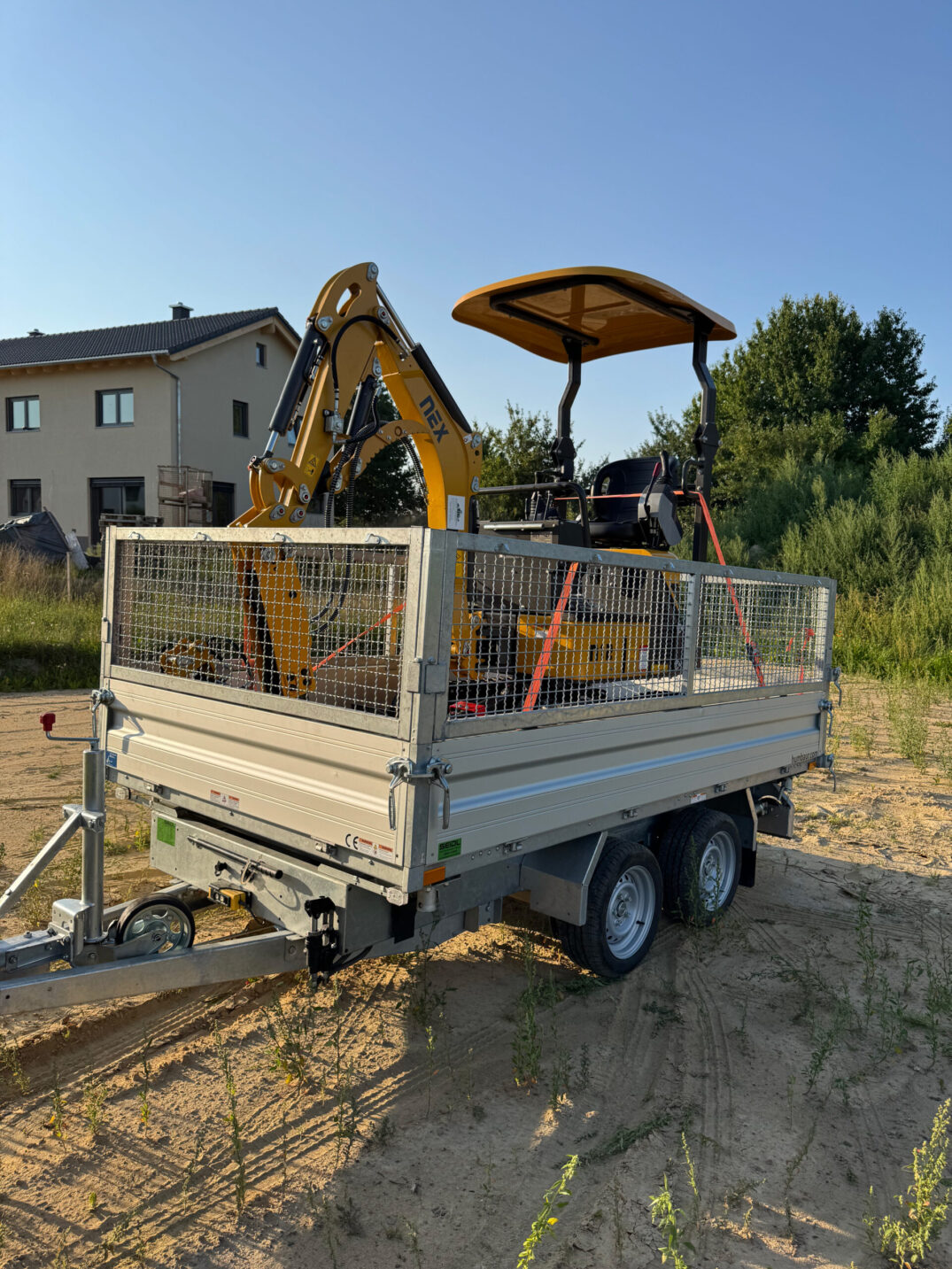 Gelber Kettenbagger mit Schutzdach, gesichert auf einem Doppelachsanhänger mit Gitteraufsätzen, geparkt auf einer sandigen Baustelle.