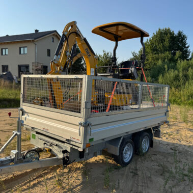 Gelber Kettenbagger mit Schutzdach, gesichert auf einem Doppelachsanhänger mit Gitteraufsätzen, geparkt auf einer sandigen Baustelle.
