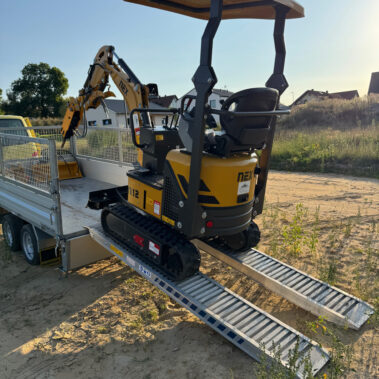 Gelber Minibagger mit Kettenantrieb fährt rückwärts über zwei Metallrampen auf einen Anhänger.