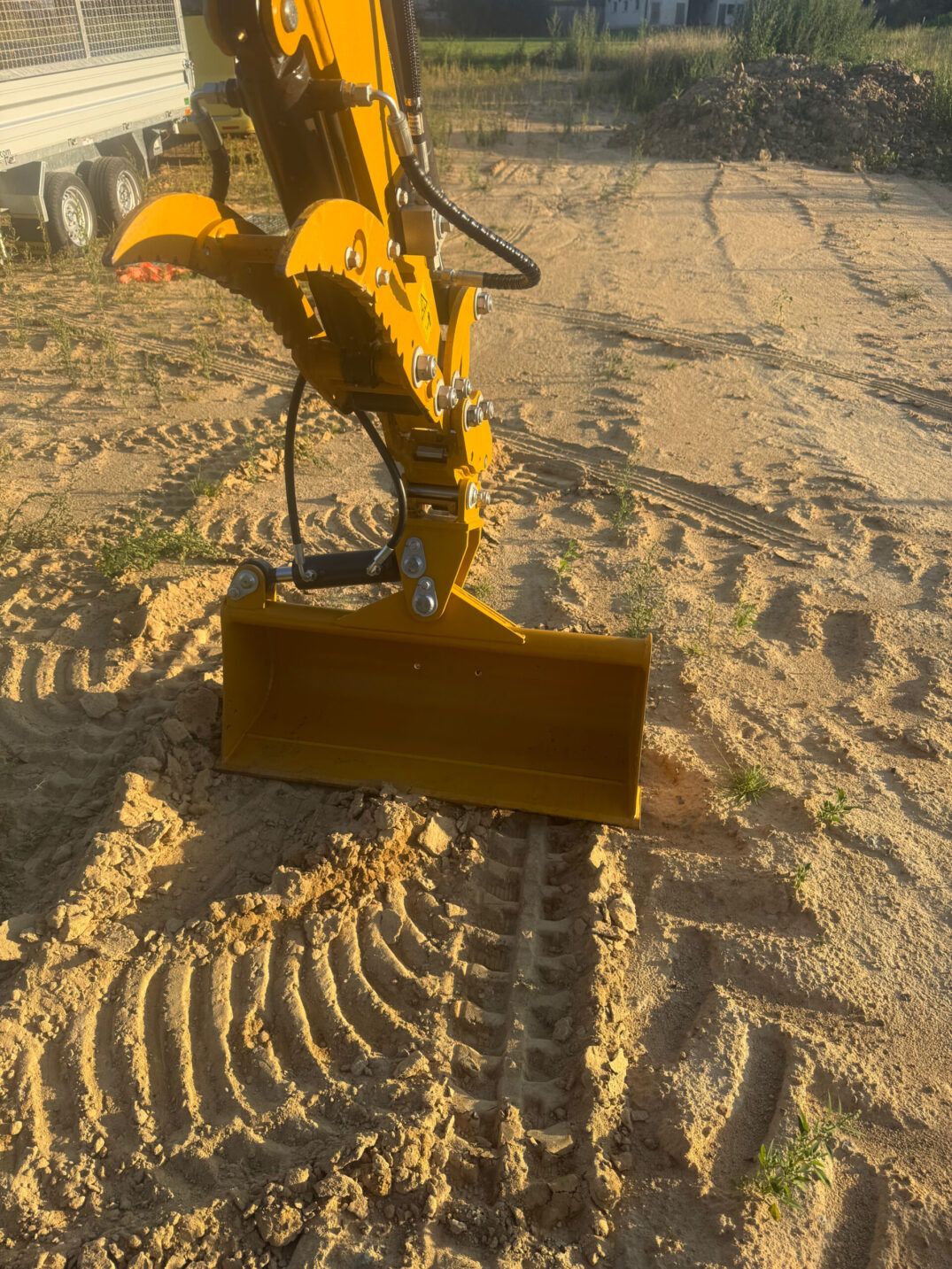 Nahaufnahme einer gelben Ladeschaufel mit groben Zähnen auf sandigem Boden, daneben Kettenabdrücke.