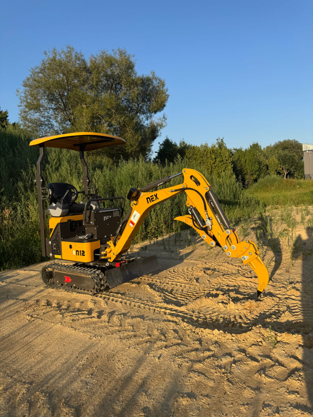 Gelber Minibagger mit montiertem Reißzahnaufsatz, auf Baustellengelände im Abendlicht.