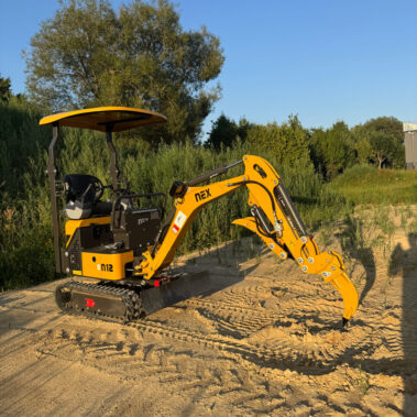 Gelber Minibagger mit montiertem Reißzahnaufsatz, auf Baustellengelände im Abendlicht.