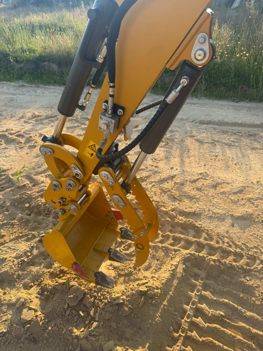 Nahaufnahme eines gelben Hydraulikarms mit montierter Schaufel, sichtbare Hydraulikleitungen und Verschraubungen.
