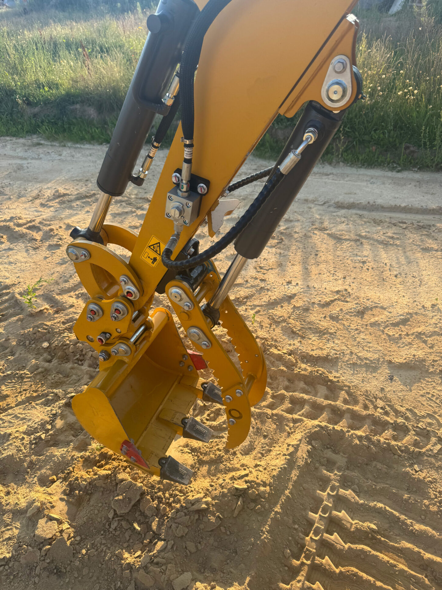 Nahaufnahme eines gelben Hydraulikarms mit montierter Schaufel, sichtbare Hydraulikleitungen und Verschraubungen.