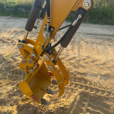 Nahaufnahme eines gelben Hydraulikarms mit montierter Schaufel, sichtbare Hydraulikleitungen und Verschraubungen.