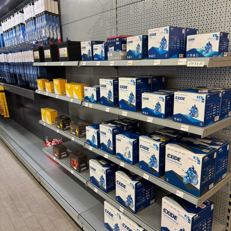 Verkaufsregal mit verschiedenen Fahrzeugbatterien in Kartonverpackungen, überwiegend blau-weiß mit Motorradabbildung.