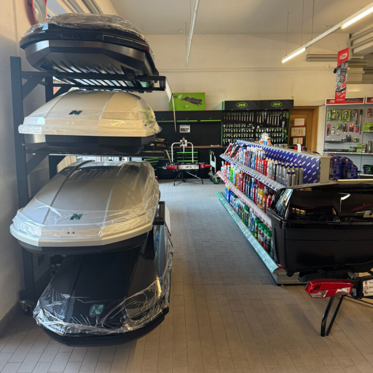 Stapel von schwarzen und weißen Dachboxen auf einem Präsentationsregal, im Hintergrund weitere Autozubehör-Artikel.