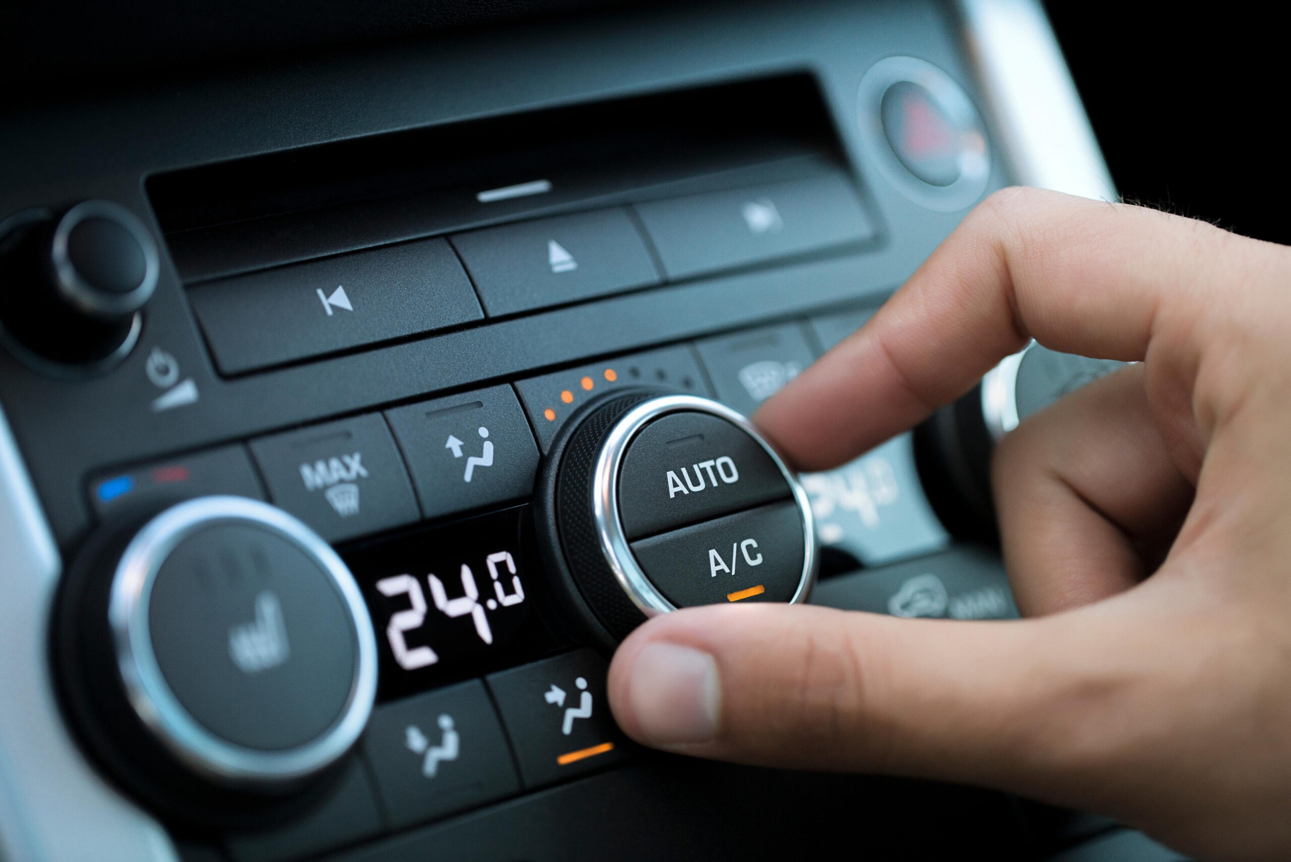 AC Auto Nahaufnahme einer Hand, die den „AUTO A/C“-Regler der Klimaanlage in einem Auto bedient. Die digitale Anzeige zeigt eine eingestellte Temperatur von 24 Grad Celsius. Die umliegenden Symbole für Lüftung, Heizung und Scheibenenteisung sind ebenfalls sichtbar.