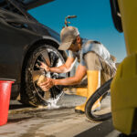 Ein Mann in Arbeitskleidung und Gummistiefeln reinigt bei Sonnenschein mit Schwamm und Seifenwasser die Felge eines dunklen Autos. Im Vordergrund stehen ein roter Eimer und ein gelber Hochdruckreiniger.