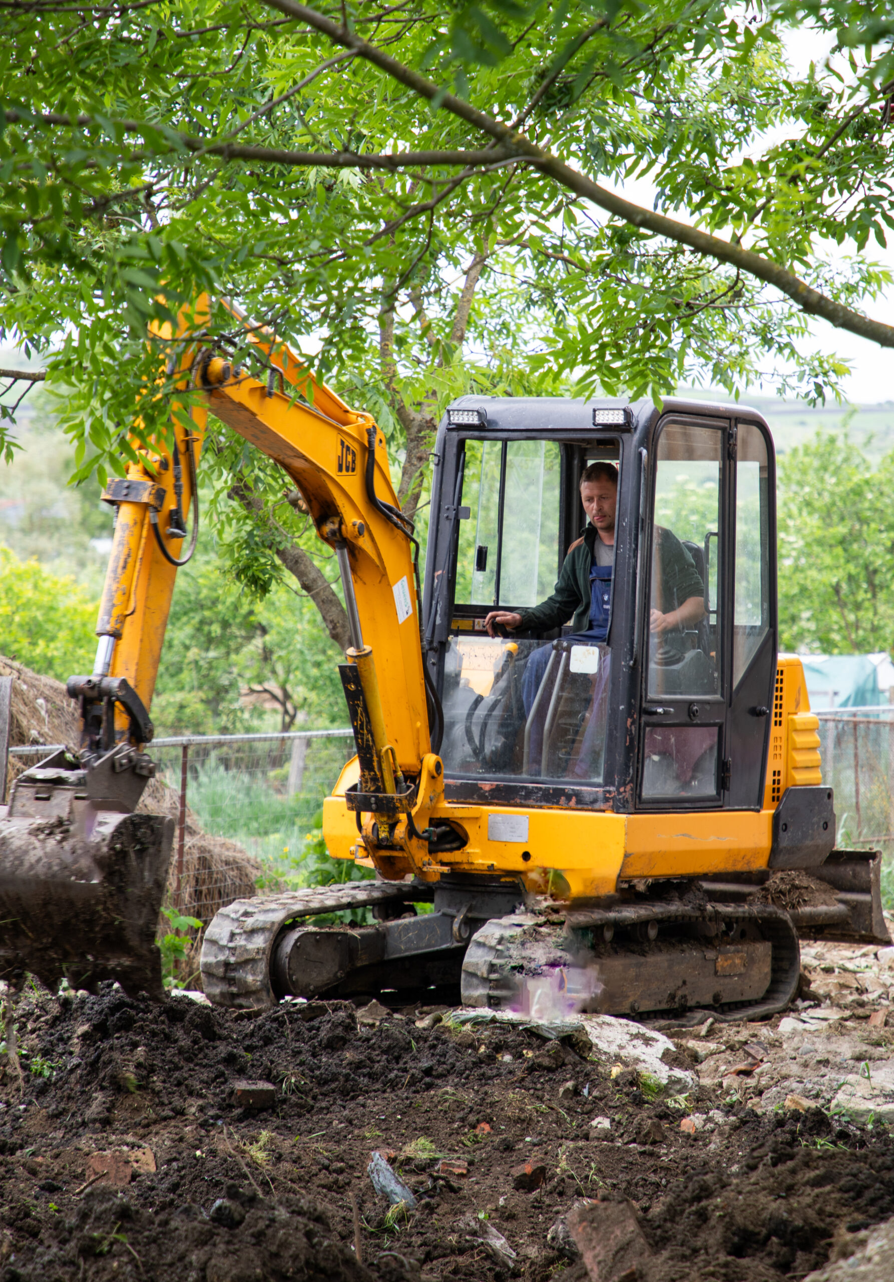 Minibagger Ein Mann bedient einen gelben Minibagger auf einer Baustelle im Grünen. Der Bagger hebt Erde mit der Schaufel aus, im Hintergrund sind Bäume und Natur zu sehen.