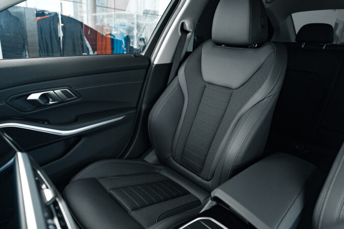 Detailansicht eines hochwertigen Vordersitzes in einem modernen Auto. Die Sitzfläche und Rückenlehne sind mit einem schwarzen, strukturierten Material bezogen und verfügen über sportliche Seitenpolster. Im Hintergrund sind leicht verschwommen Kleidungsstücke in einem Schaufenster zu erkennen. Das Interieur wirkt hochwertig und gepflegt.