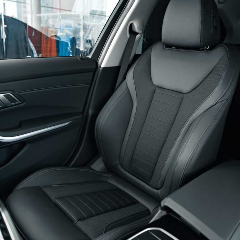 Detailansicht eines hochwertigen Vordersitzes in einem modernen Auto. Die Sitzfläche und Rückenlehne sind mit einem schwarzen, strukturierten Material bezogen und verfügen über sportliche Seitenpolster. Im Hintergrund sind leicht verschwommen Kleidungsstücke in einem Schaufenster zu erkennen. Das Interieur wirkt hochwertig und gepflegt.