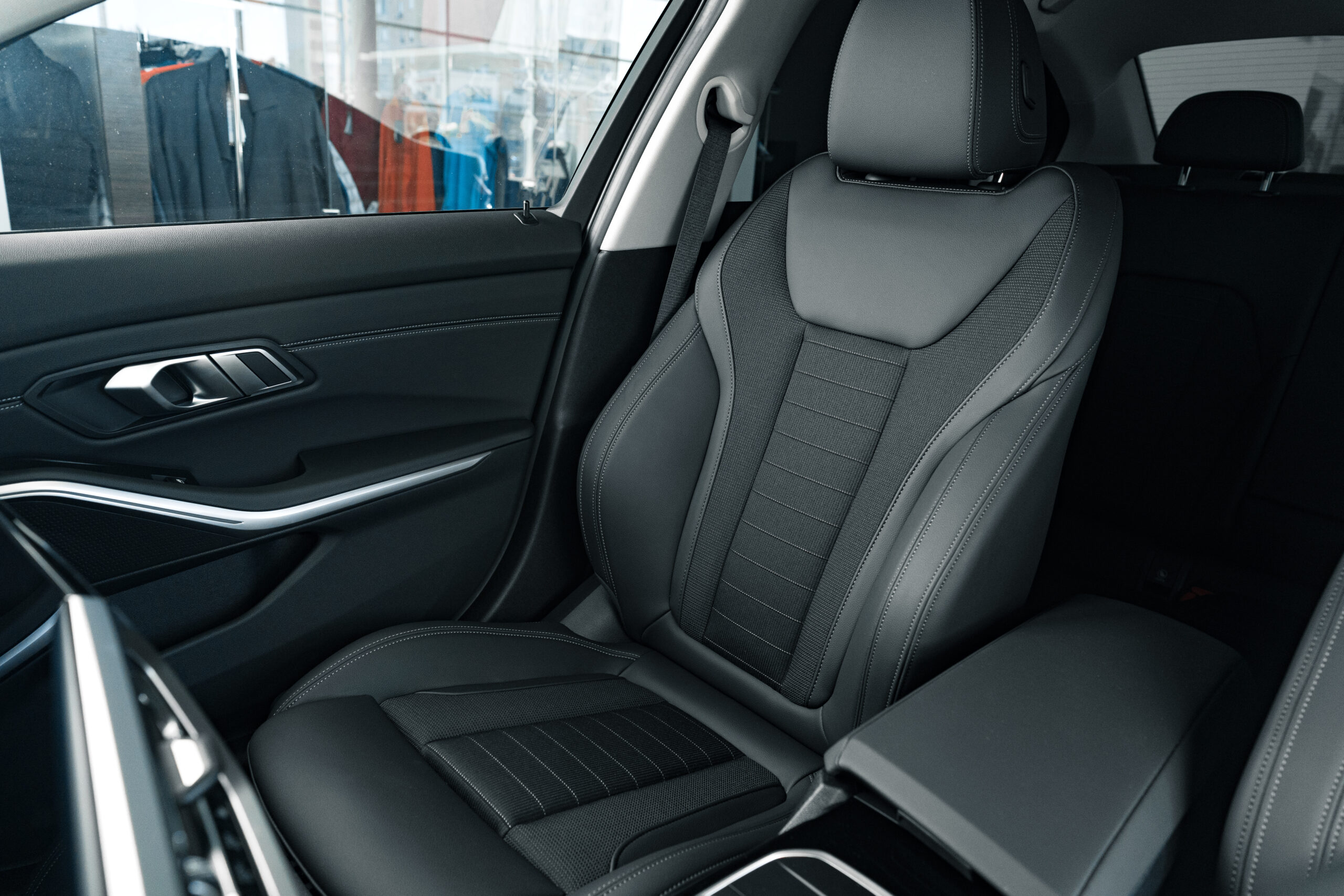 Sitzbezüge Detailansicht eines hochwertigen Vordersitzes in einem modernen Auto. Die Sitzfläche und Rückenlehne sind mit einem schwarzen, strukturierten Material bezogen und verfügen über sportliche Seitenpolster. Im Hintergrund sind leicht verschwommen Kleidungsstücke in einem Schaufenster zu erkennen. Das Interieur wirkt hochwertig und gepflegt.