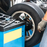 Ein Werkstattmitarbeiter mit Handschuhen steht an einer Auswuchtmaschine, auf der gerade ein Autoreifen rotiert. Auf der Maschine liegen verschiedene Werkzeuge und Gewichte, im Hintergrund sind weitere Arbeitsmaterialien zu sehen.