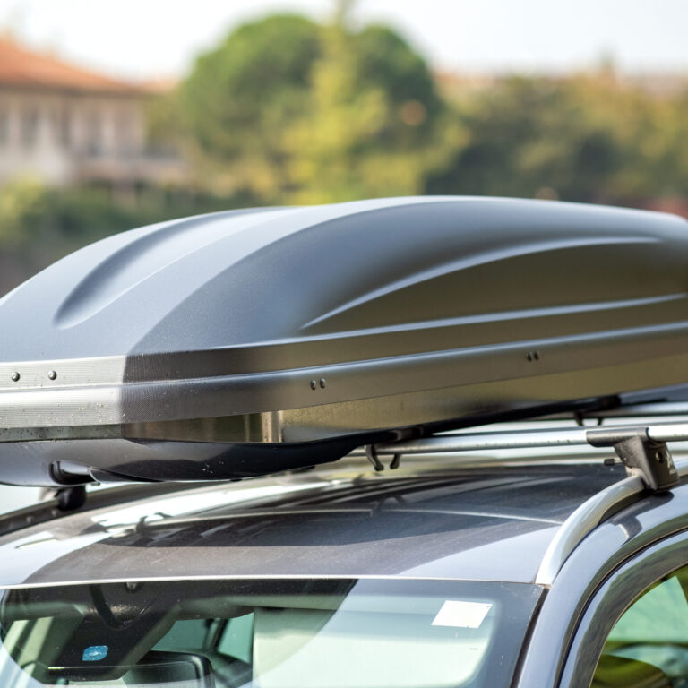Auto mit montierter schwarzer Dachbox auf einem Dachträger, im Hintergrund eine unscharfe Fluss- und Häuserkulisse.
