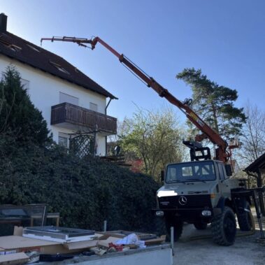LKW mit Kran hebt Material auf das Dach eines Einfamilienhauses, Bauarbeiten im Gartenbereich.