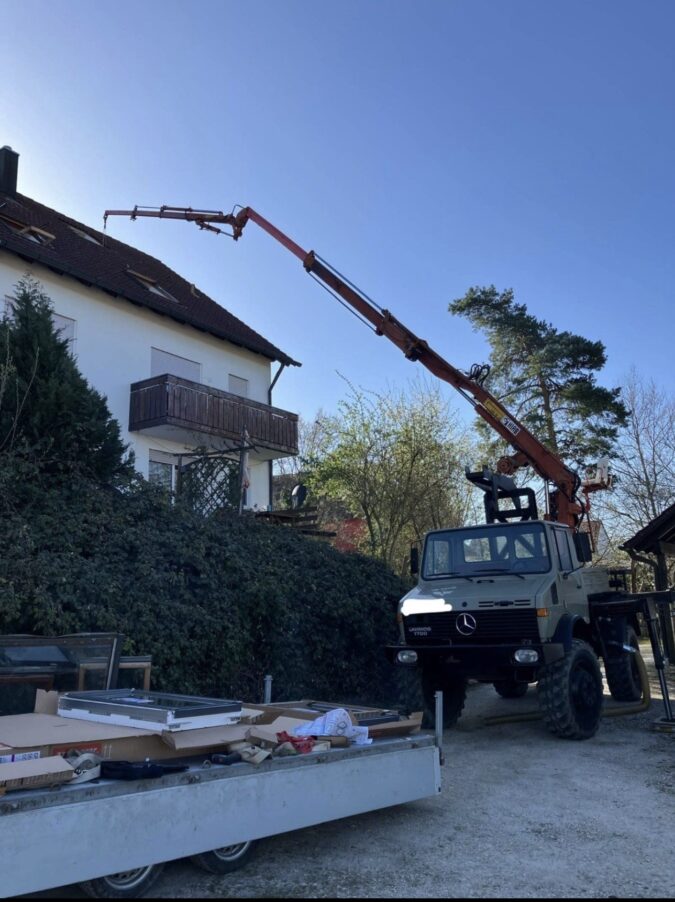 LKW mit Kran hebt Material auf das Dach eines Einfamilienhauses, Bauarbeiten im Gartenbereich.