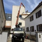 Kran hebt lange Holzbalken über ein Gebäude hinweg in einen Innenhof zwischen mehreren Häusern.