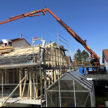 Kran montiert Holzelemente auf ein Haus im Bau, Arbeiter stehen auf dem Dachgerüst.