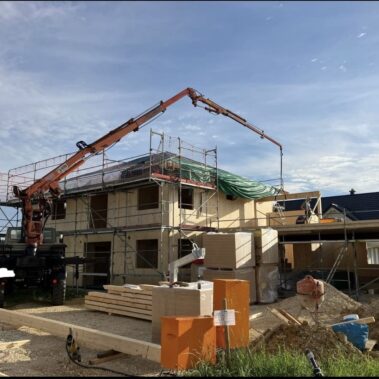 Baustelle mit Holzhaus im Aufbau, Kran hebt Bauteile auf das Gerüstdach.
