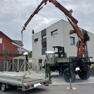 Kran hebt lange Bauteile während Montage eines Carports vor einem Wohnhaus.