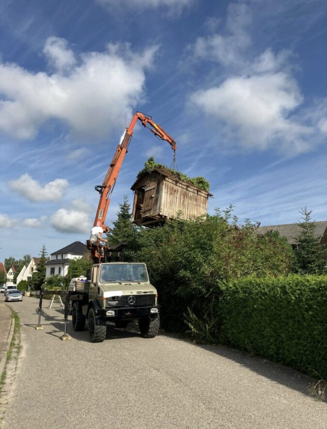 Kran hebt ein kleines Holzhäuschen über eine Hecke in einen Garten.